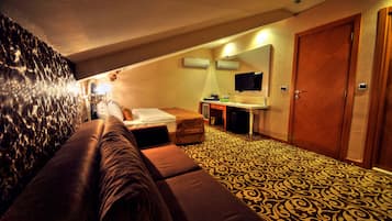 A hotel room with a patterned carpet, a sofa, a bed, a desk with a computer, a TV, and a wooden door.