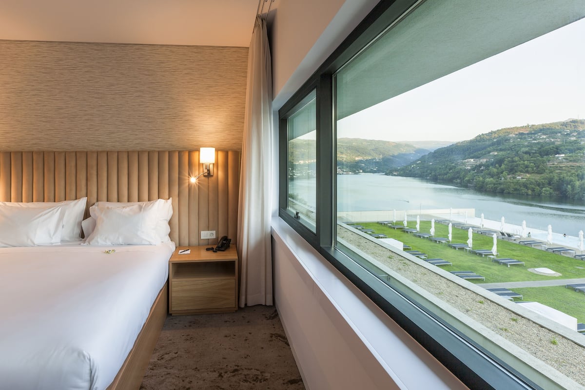 Une chambre d’hôtel avec un grand lit, des lampes de chevet, une table de nuit et une vue sur une rivière et des collines.