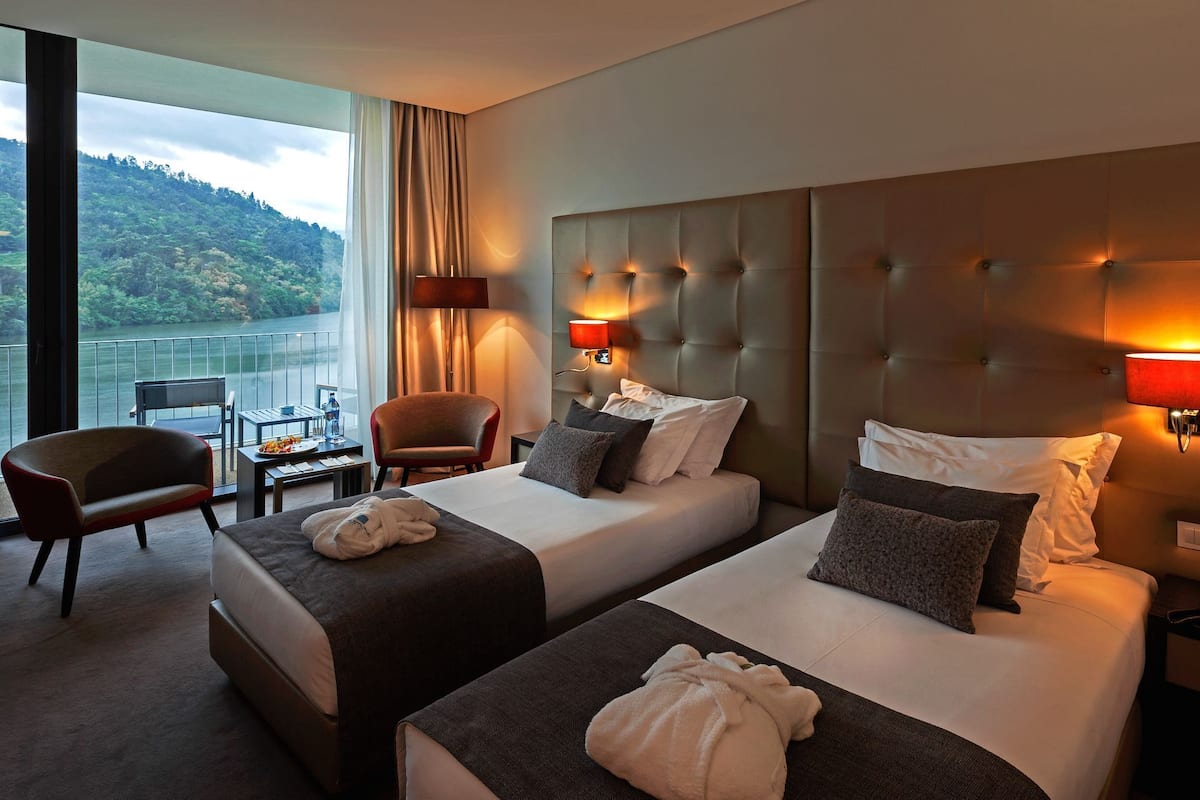 Une chambre d’hôtel avec deux lits, un coin salon comprenant un fauteuil et une petite table, et offrant une vue sur un lac et des montagnes.