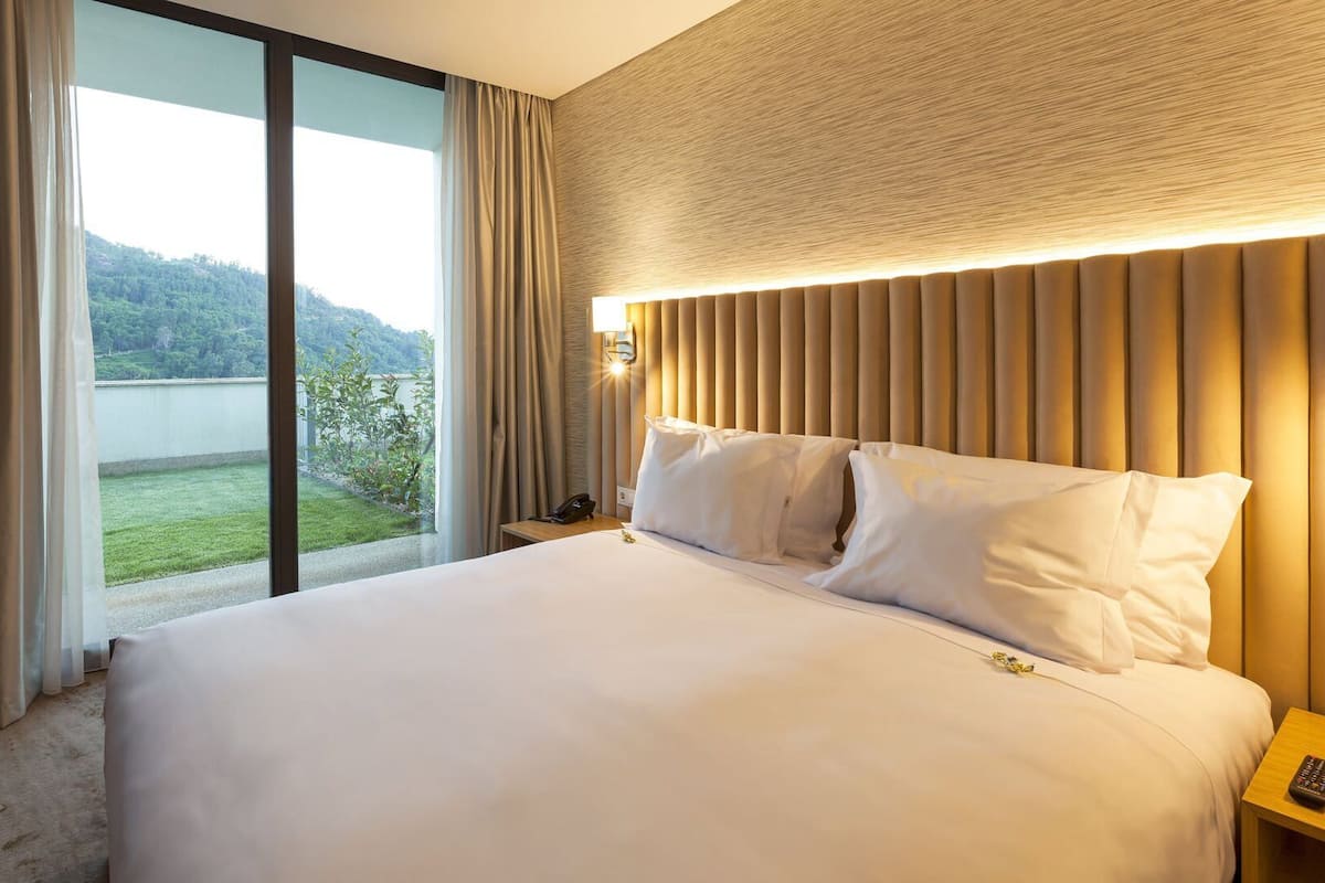Une chambre d’hôtel avec un grand lit, une table de chevet avec une lampe et une vue sur un paysage verdoyant, visible à travers une porte-fenêtre coulissante.