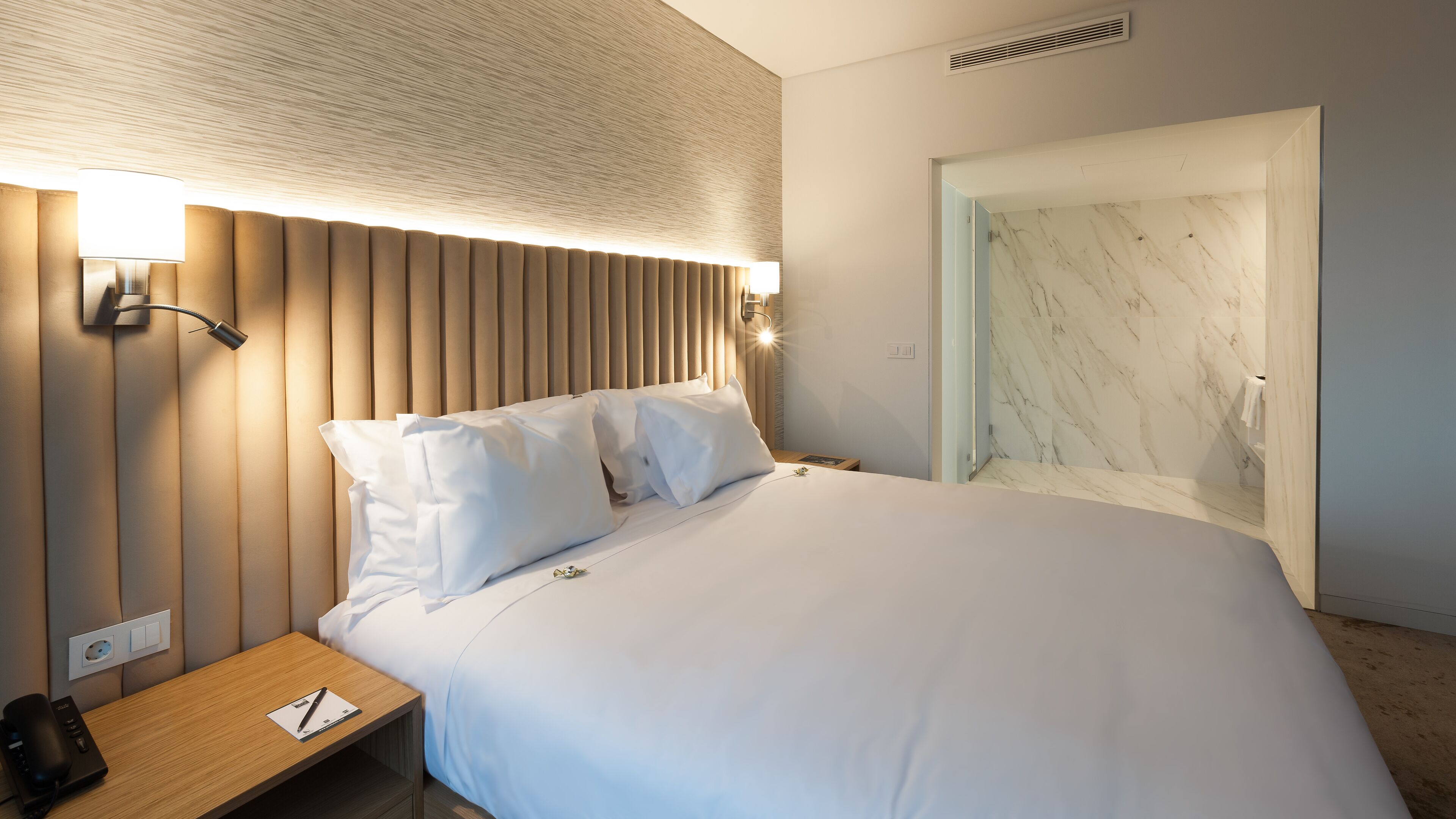 A modern hotel room with a large bed, a wooden nightstand, a lamp, and a marble wall.