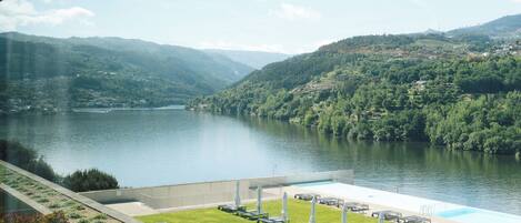Vista de un lago con montañas al fondo, piscina con sillones de descanso y una zona verde.