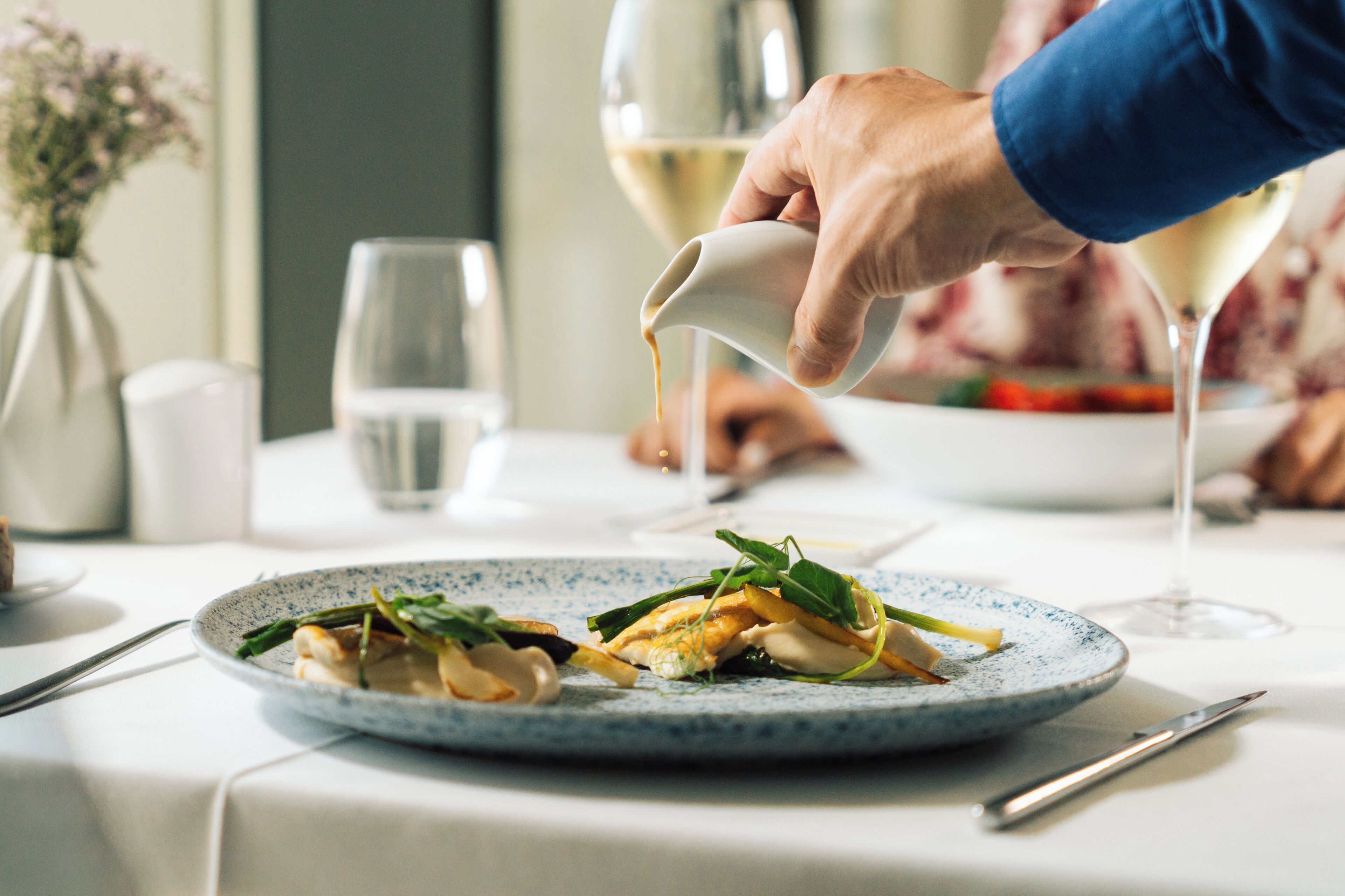 Una mesa de comedor con un plato de comida, una copa de vino y un jarrón con flores.