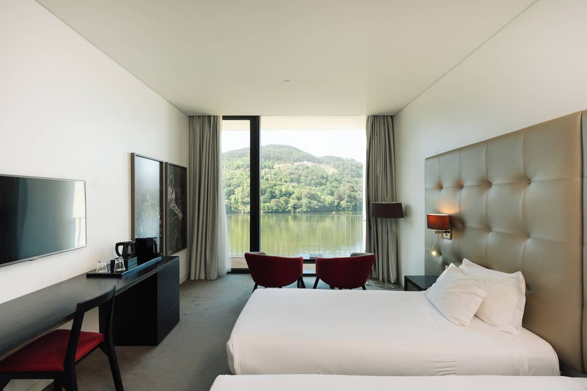 Une chambre d’hôtel comprenant un lit, un bureau avec une télévision, deux chaises, une fenêtre donnant sur un lac et des collines, et une lampe fixée au mur.