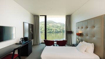 Habitación de hotel con cama, escritorio con televisor, dos sillas, ventana con vista a un lago y colinas, y una lámpara en la pared.