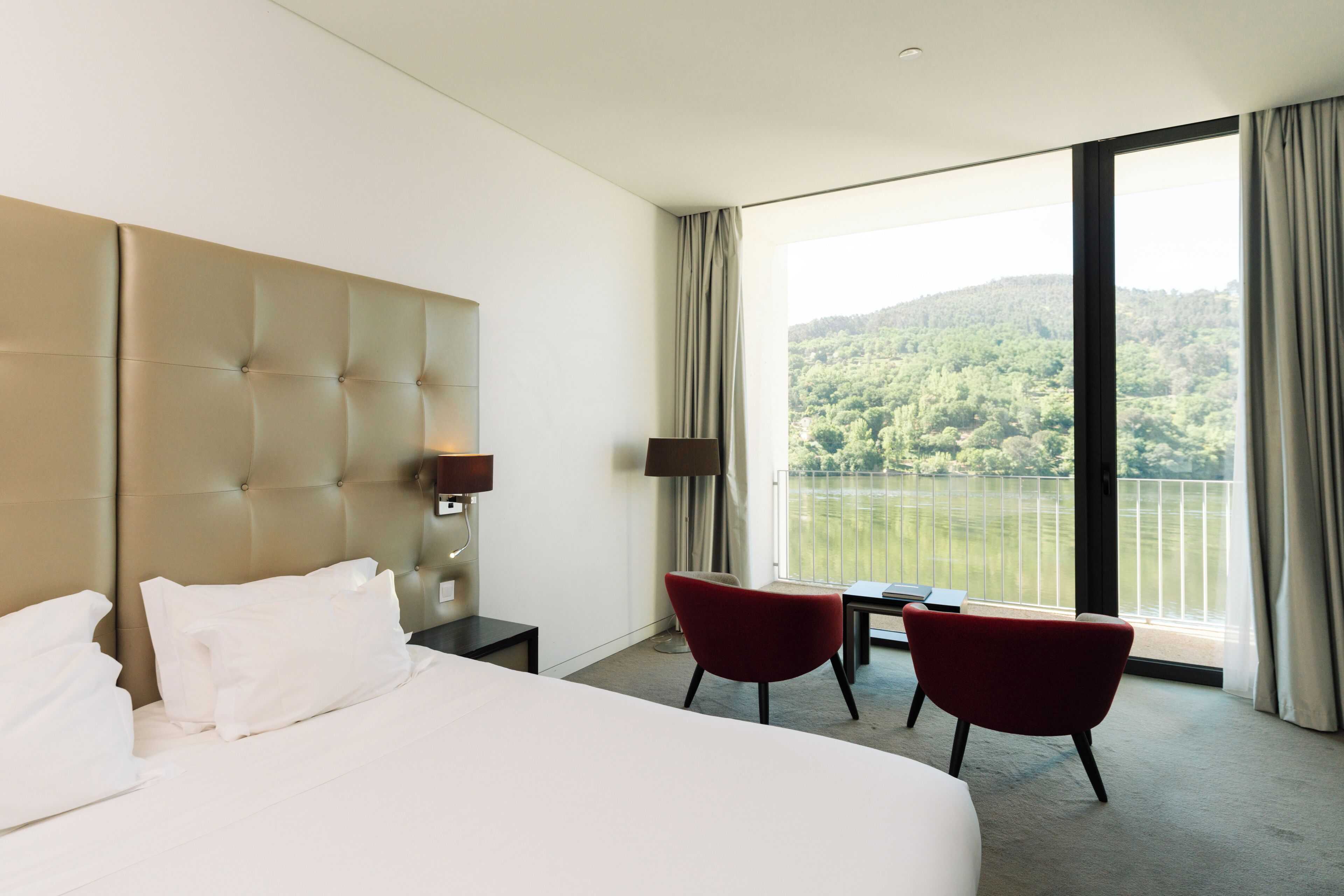 A hotel room with a large bed, two chairs, a small table, a lamp, and a large window with a view of a lake and forest.