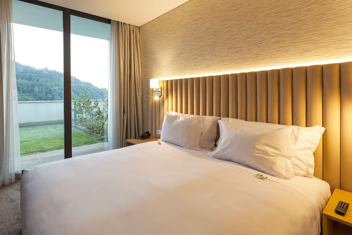 Une chambre d’hôtel avec un grand lit, une table de chevet avec une lampe et une vue sur un paysage verdoyant par la fenêtre.