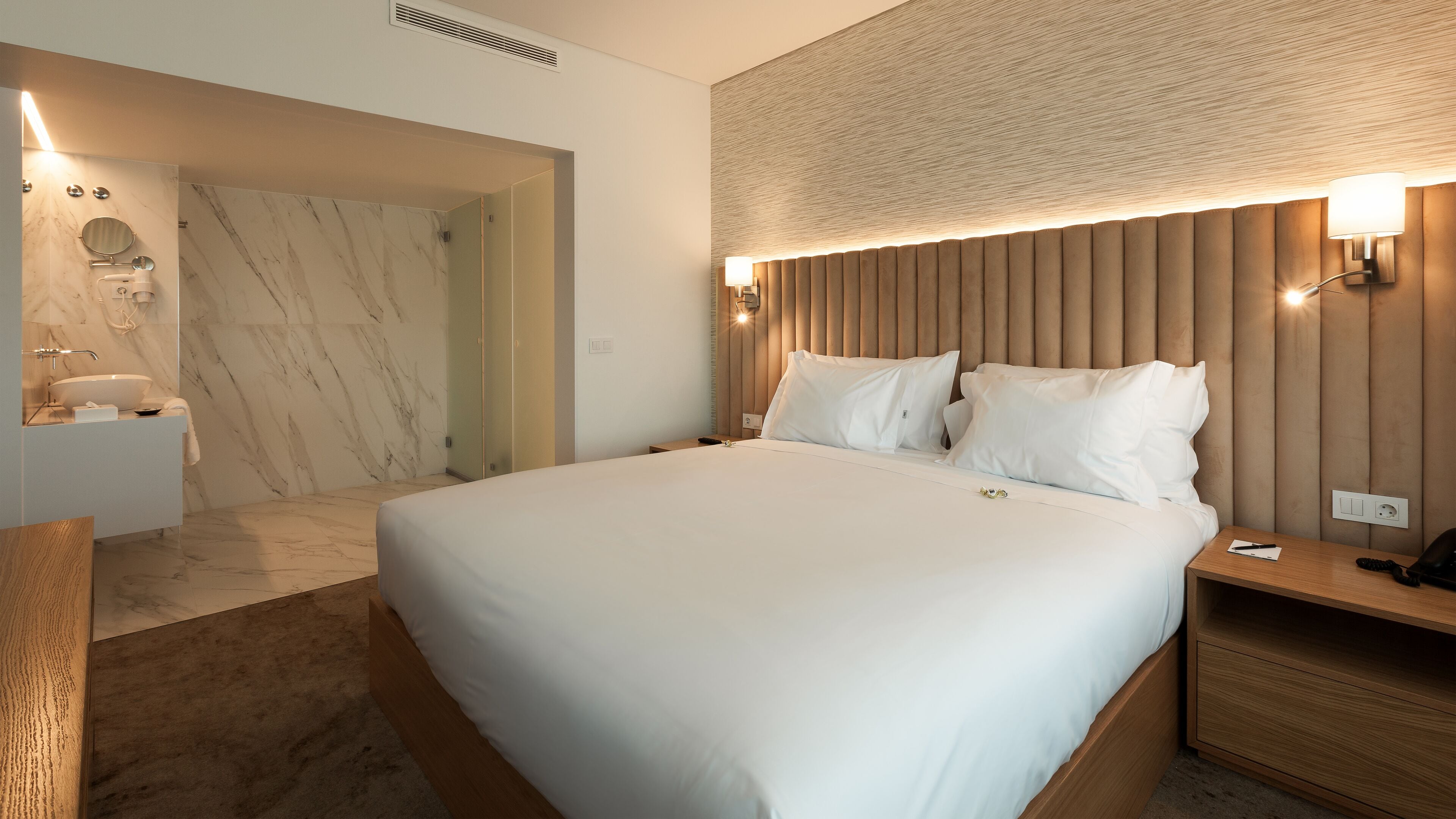 A modern hotel room with a large bed, a wooden headboard with wall-mounted lights, a marble bathroom with a round mirror, and a wooden nightstand.