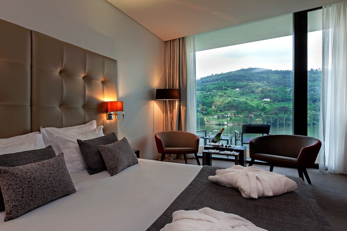 Une chambre d’hôtel dotée d’un grand lit, offrant une vue sur un paysage verdoyant, comprenant un coin salon avec une table et des chaises, et un balcon avec une balustrade.