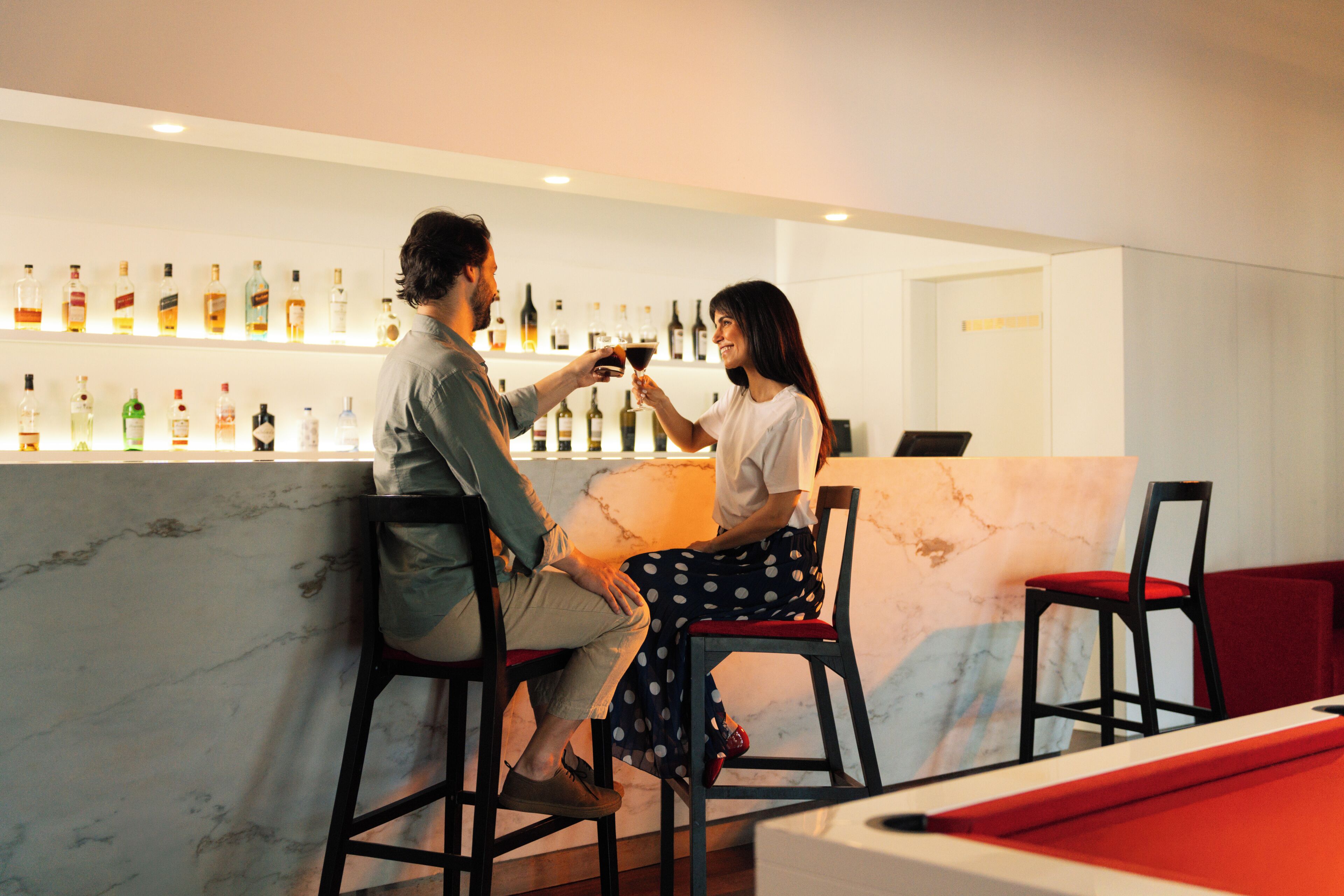 Eine Barbereitschaft mit einer Marmorplatte und mehreren alkoholischen Getränken. Im Vordergrund befinden sich zwei Barhocker und ein Billardtisch.
