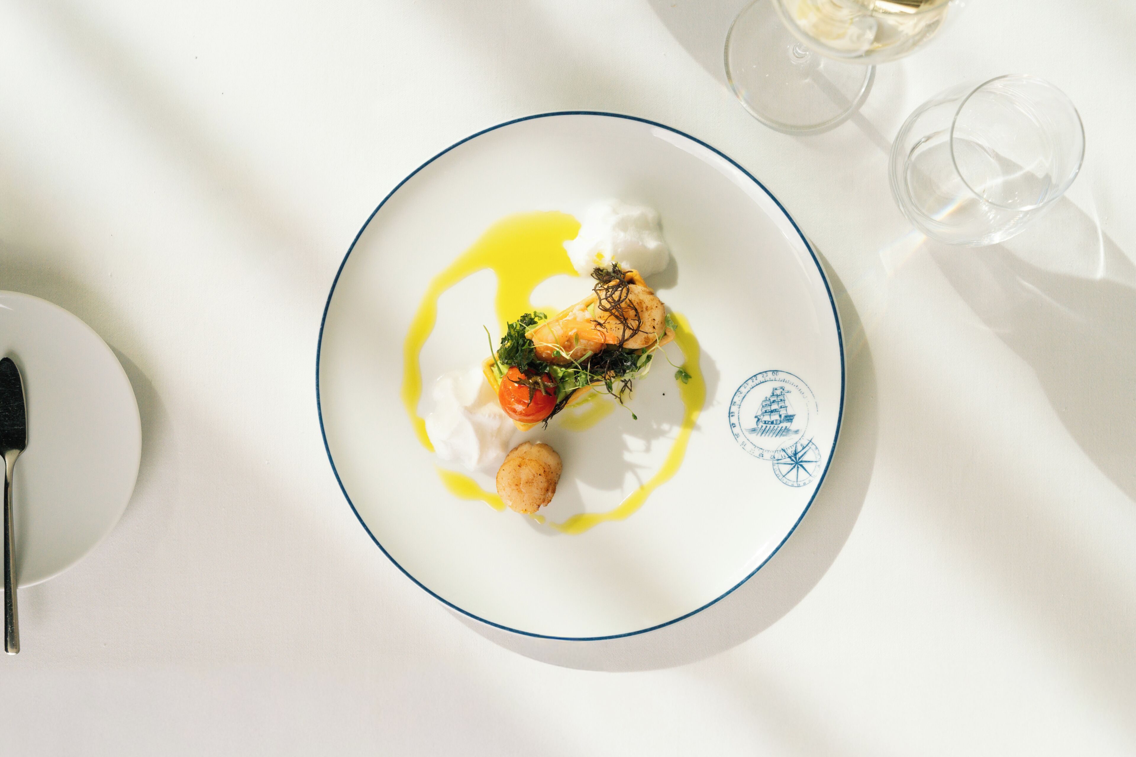 Un plato blanco con borde azul, un tenedor y dos copas de vino blanco sobre una mesa.