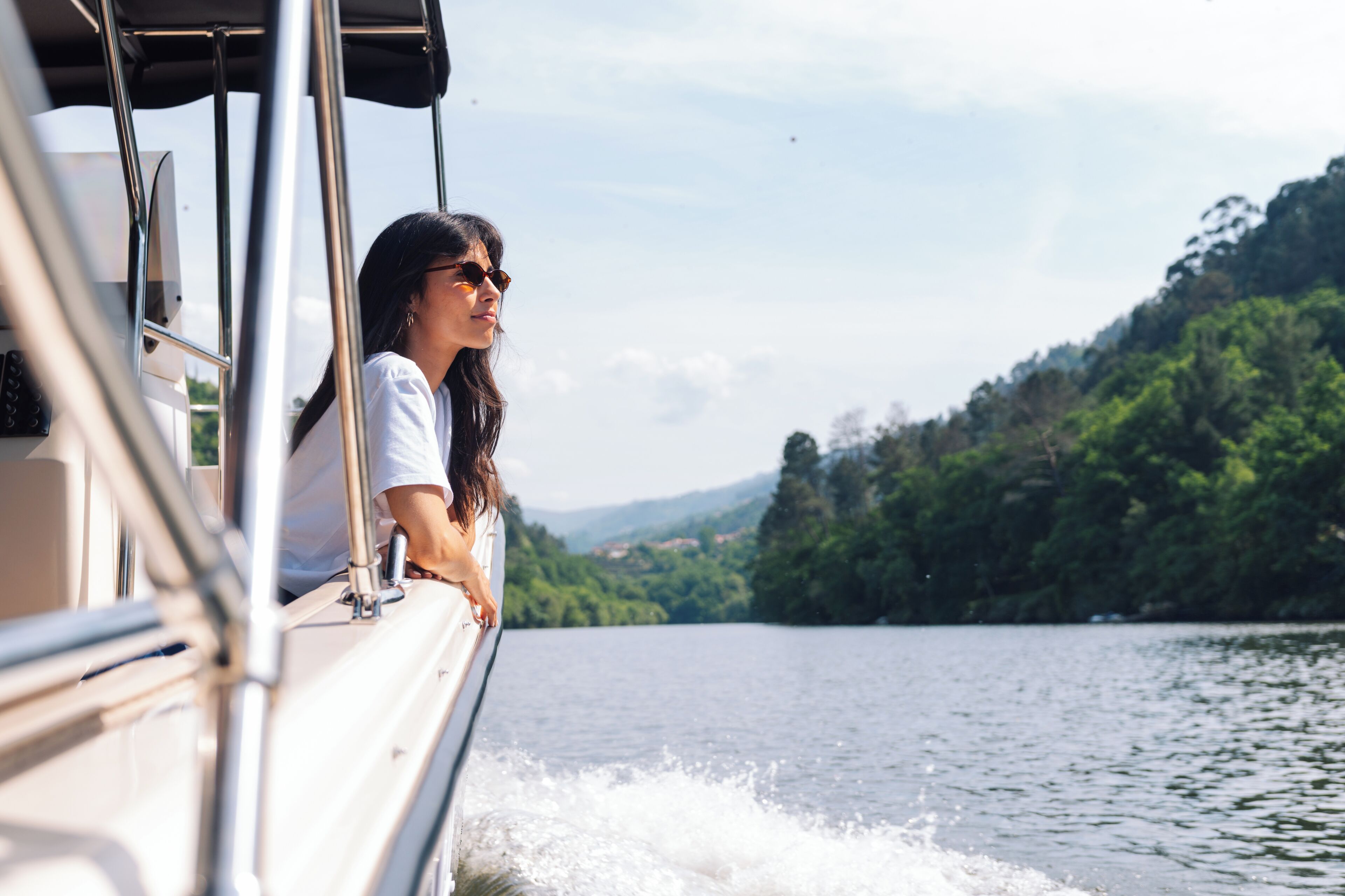 Un bote con una persona apoyada en la barandilla, un lago y una ladera boscosa al fondo.