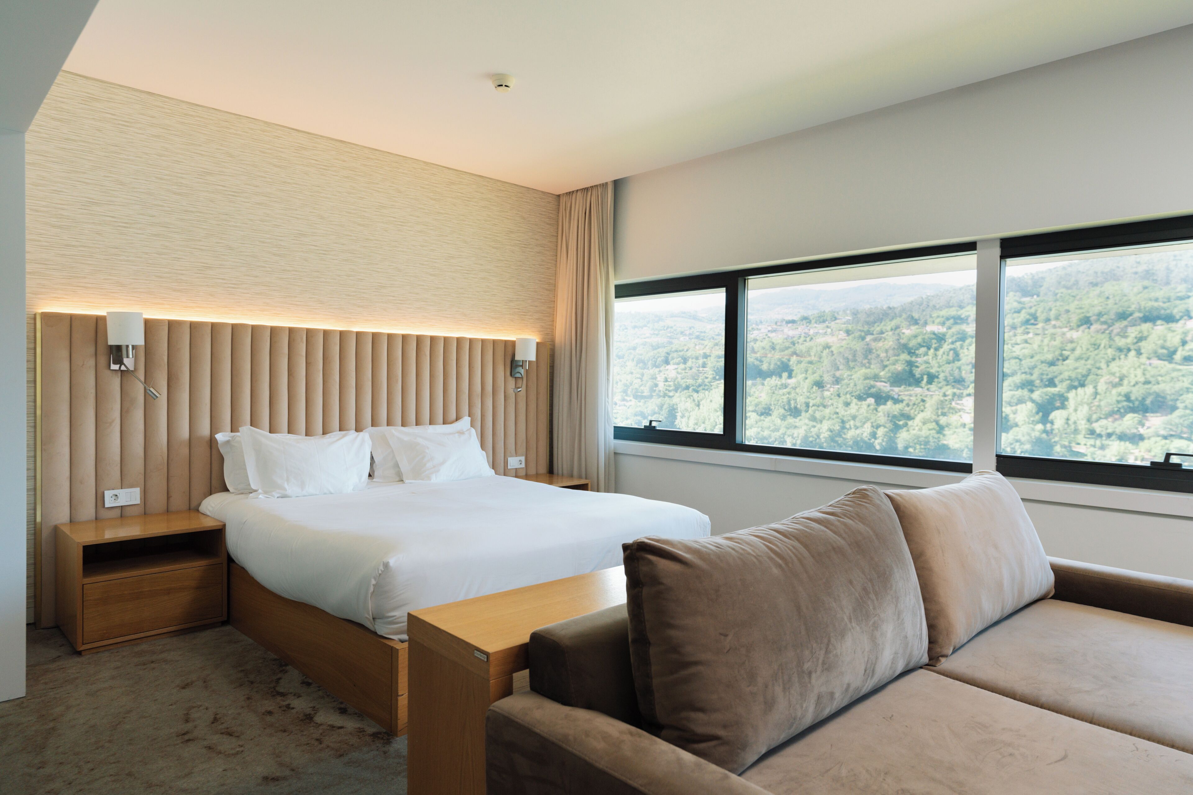 Habitación de hotel moderna con una cama grande, una mesita de noche de madera, un pequeño escritorio, un sofá y un ventanal con vistas a un bosque.