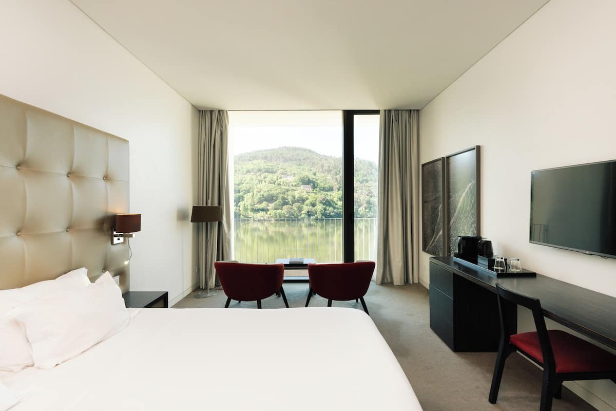 Une chambre d’hôtel comprenant un lit, un bureau, deux chaises, une télévision et une grande fenêtre donnant sur un lac et des collines.