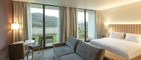 A hotel room with a bed, a sofa, a table, and chairs. There is a view of greenery outside the window.