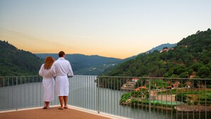 A couple standing on a balcony overlooking a river with a view of a town and hills. - Douro Royal Valley Hotel & SPA (Baiao)