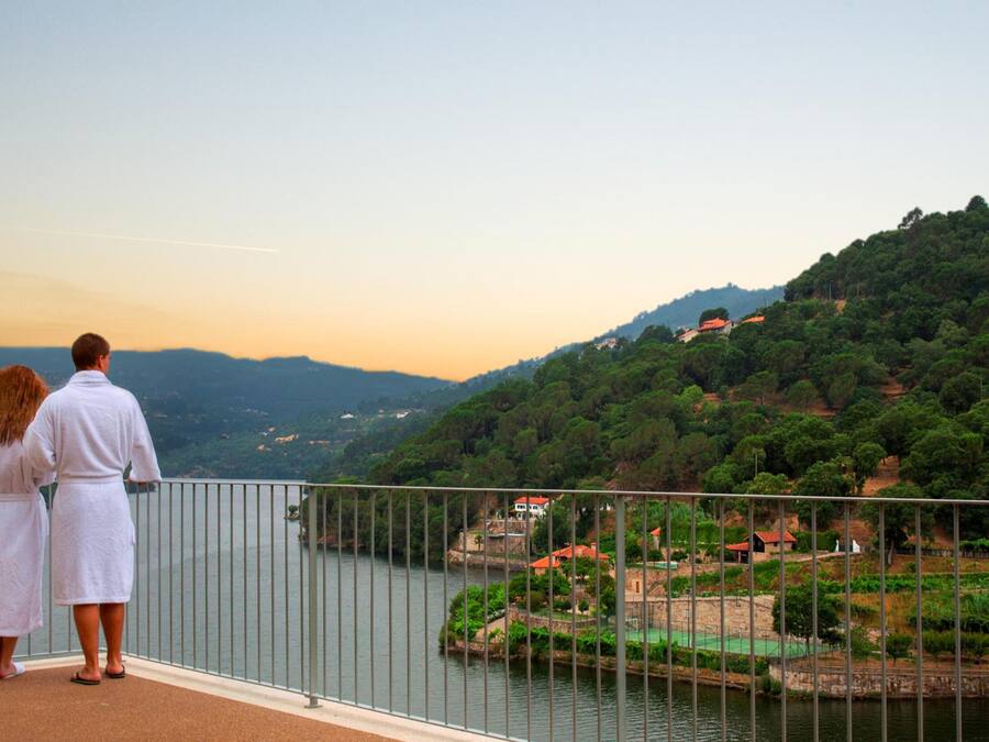 A couple standing on a balcony overlooking a river with a view of a town and hills.