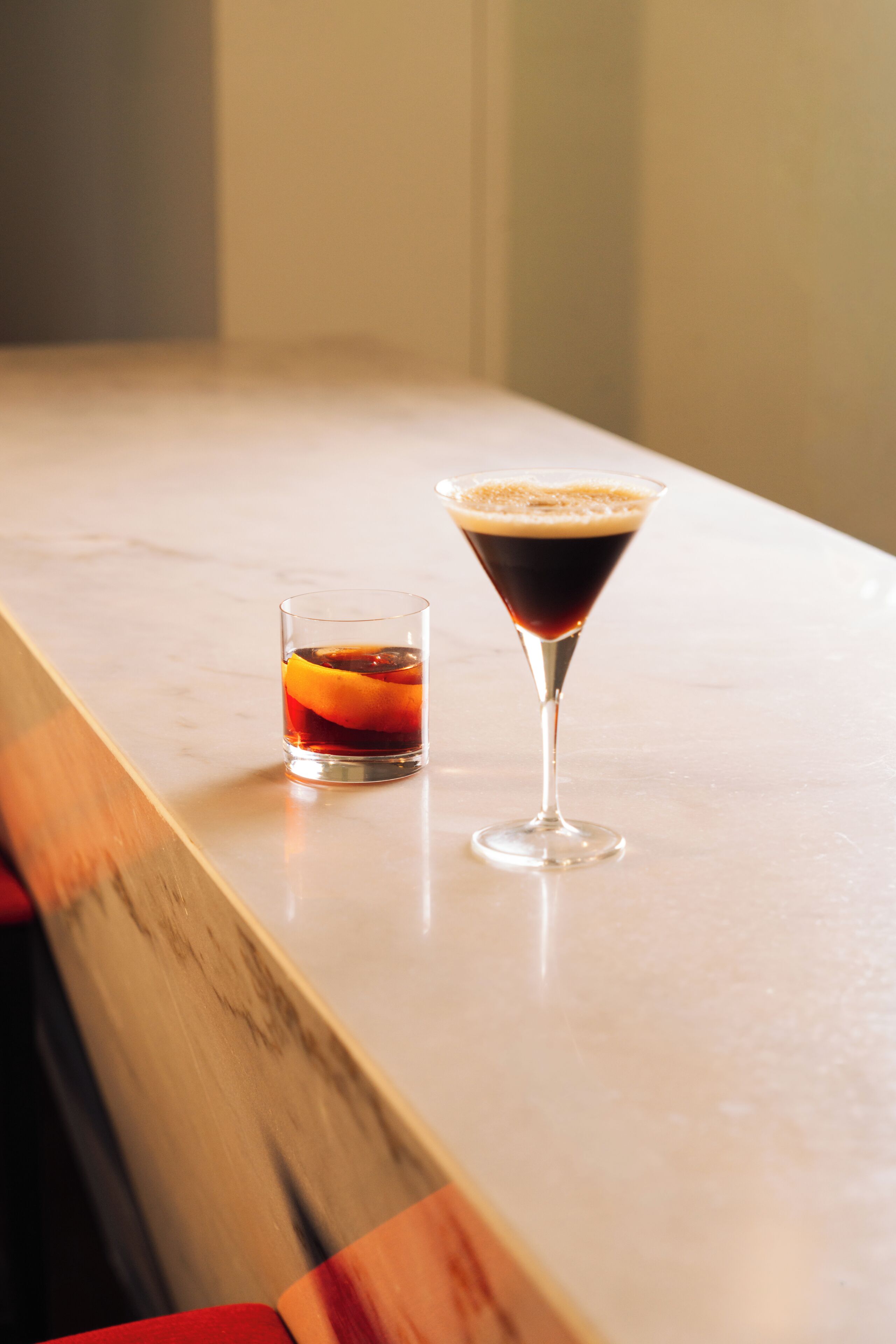 A bar area with two glasses of a dark beverage, one in a short glass and the other in a martini glass, on a marble countertop.