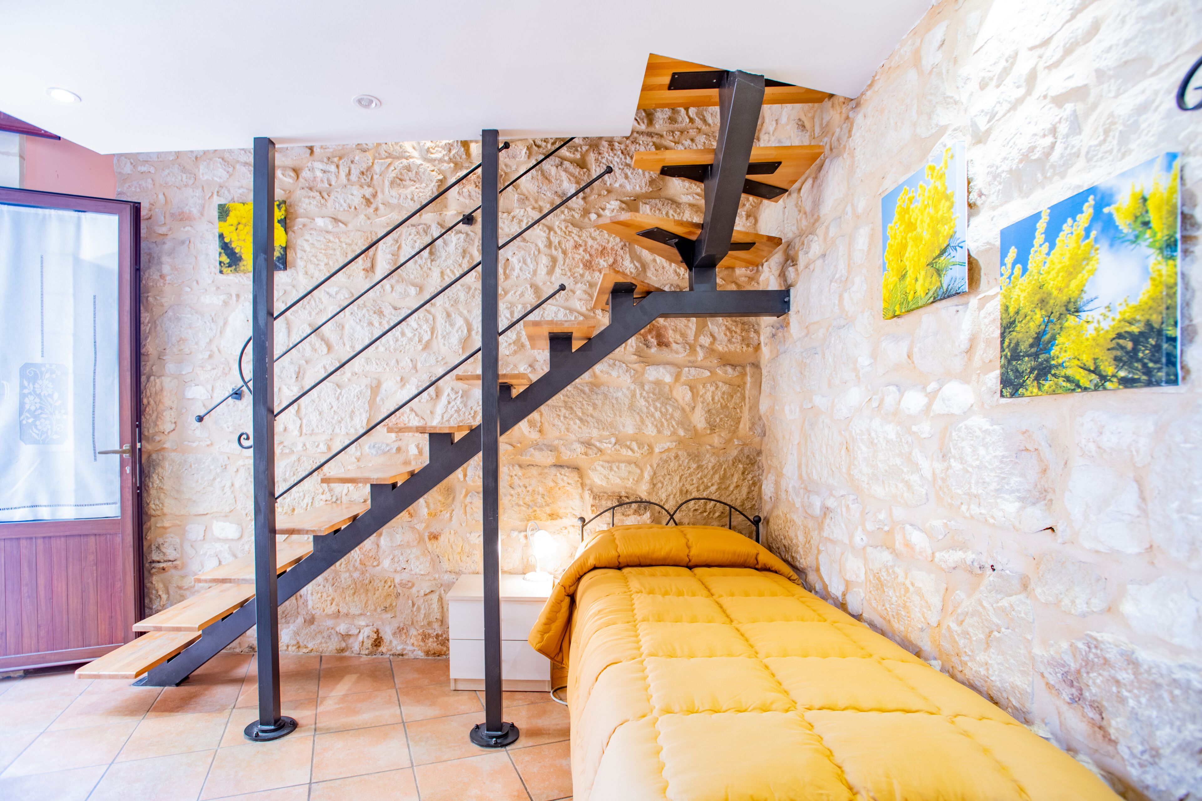 A bedroom with a stone wall, a wooden staircase, a bed with a yellow duvet, and two paintings of yellow flowers.