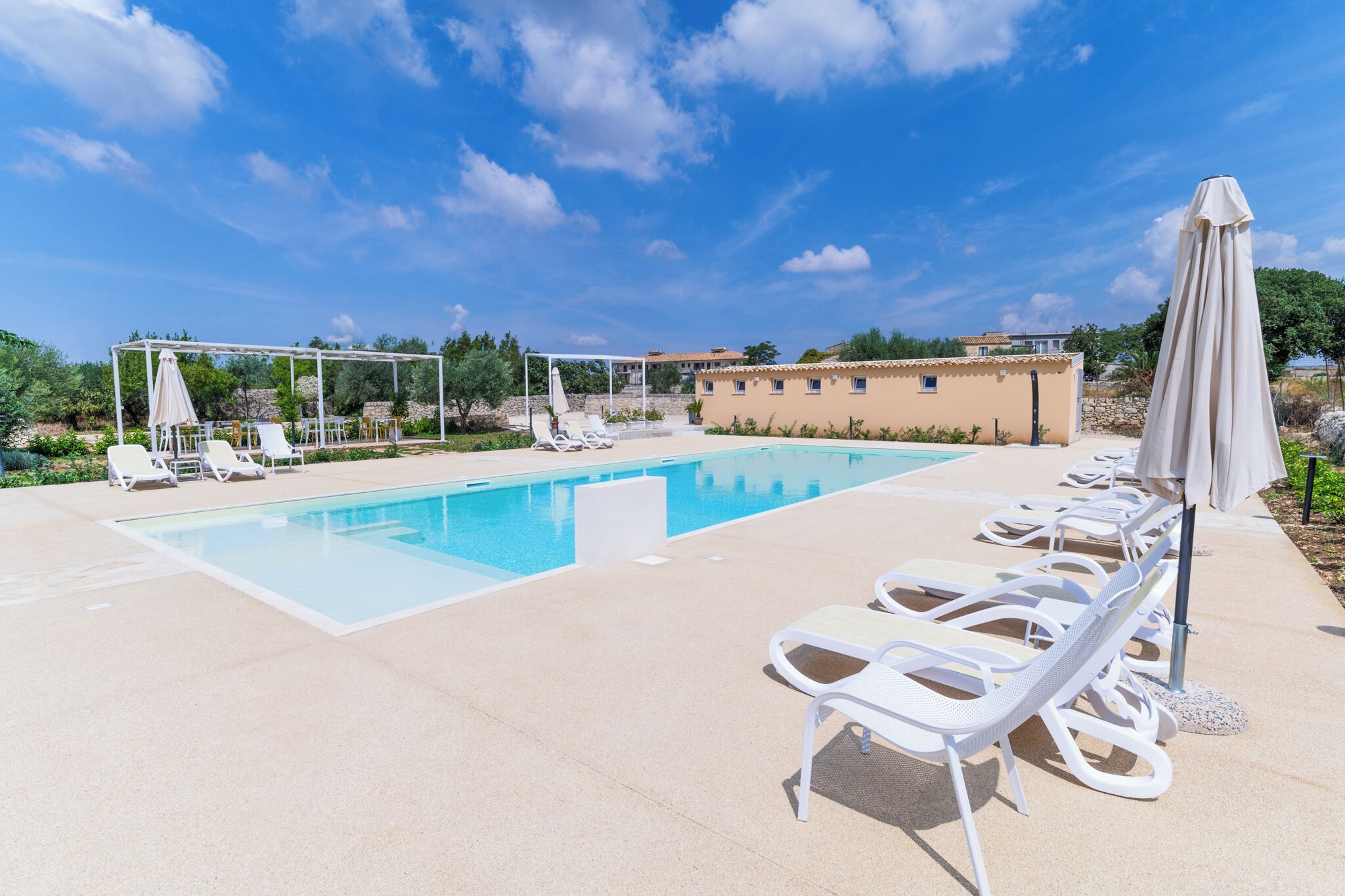A pool area with lounge chairs and umbrellas, a pergola, and a building in the background.