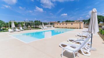 A pool area with lounge chairs and umbrellas, a pergola, and a building in the background.