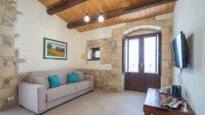 A living room with a stone wall, a wooden ceiling, a beige sofa with teal cushions, a wooden coffee table, and a television mounted on the wall. - Borgo Monachella (Ragusa)