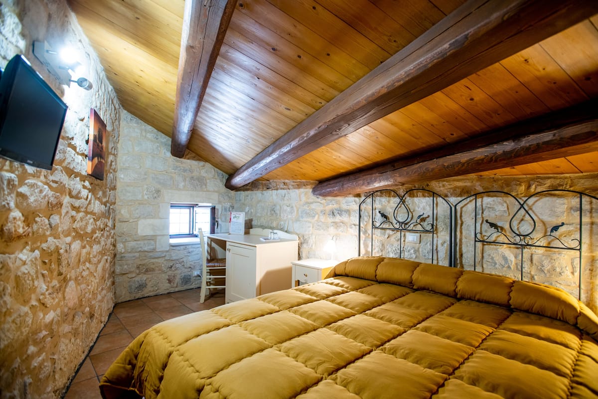 A cozy bedroom with a wooden ceiling, stone walls, a bed with a yellow duvet, a desk, and a window.