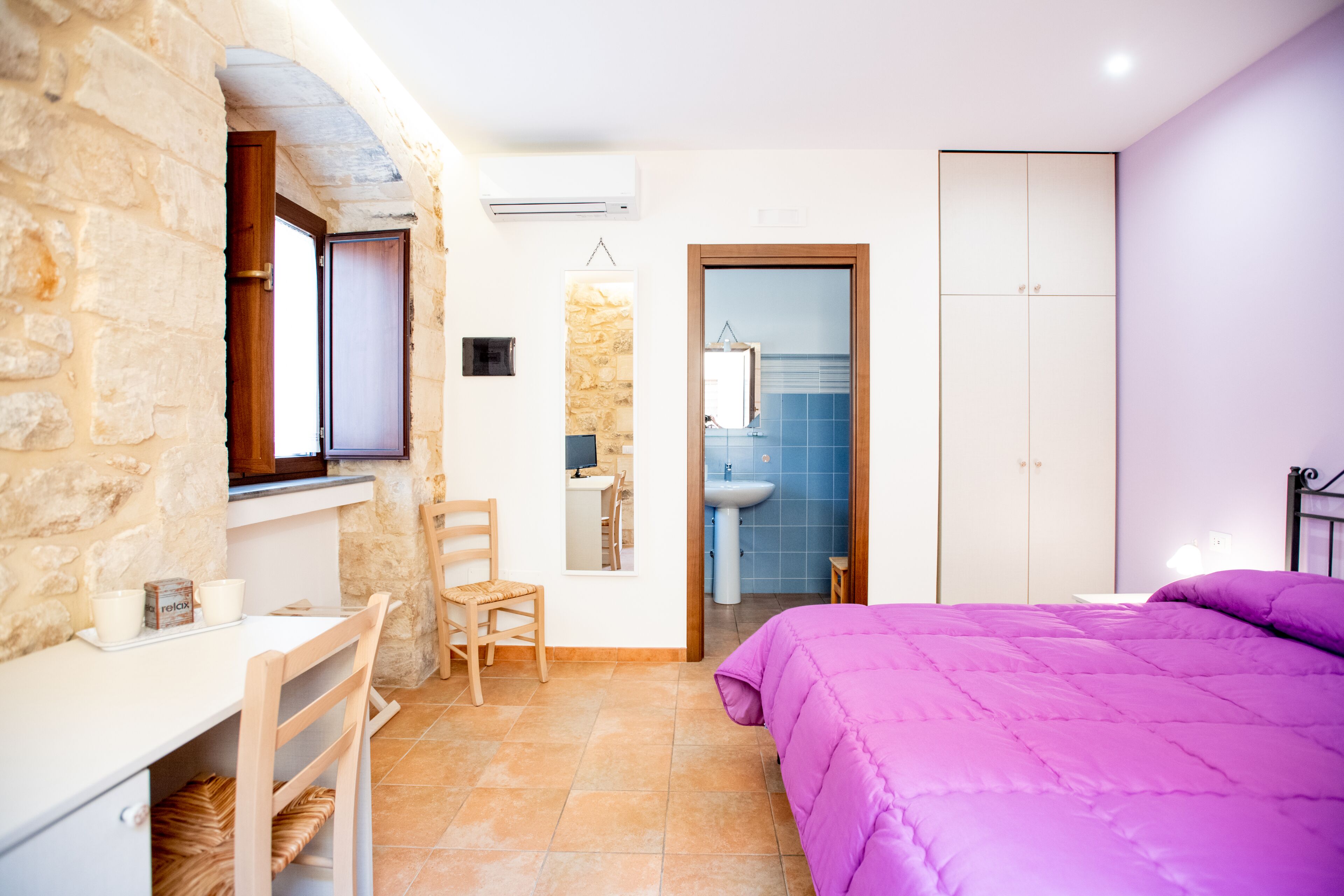 A bedroom with a stone wall, a wooden desk, a chair, a bed with a purple bedspread, and a wardrobe.