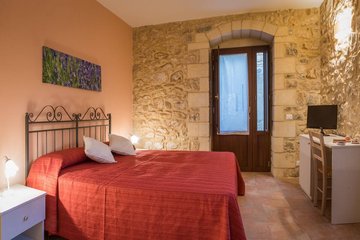 A bedroom with a stone wall, a bed with a red bedspread, a nightstand with a lamp, and a desk with a computer.