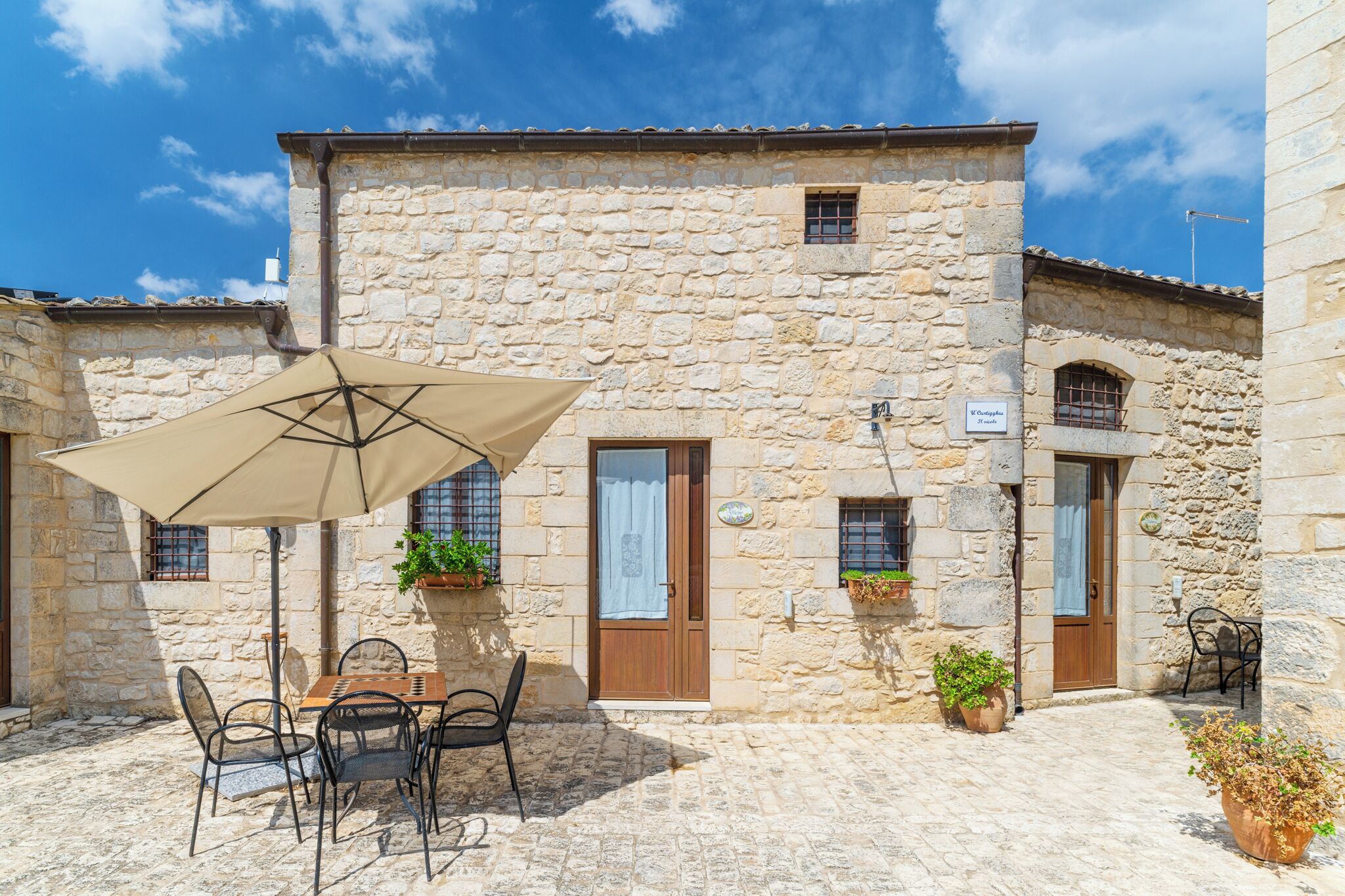 Un bâtiment en pierre doté d’une cour, d’un espace extérieur aménagé avec une table et des chaises, et de plantes en pot.