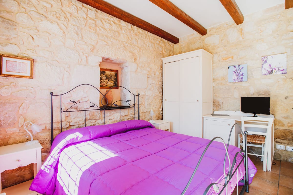 A bedroom with a stone wall, a bed with a purple bedspread, a nightstand, a desk with a chair, and a television.