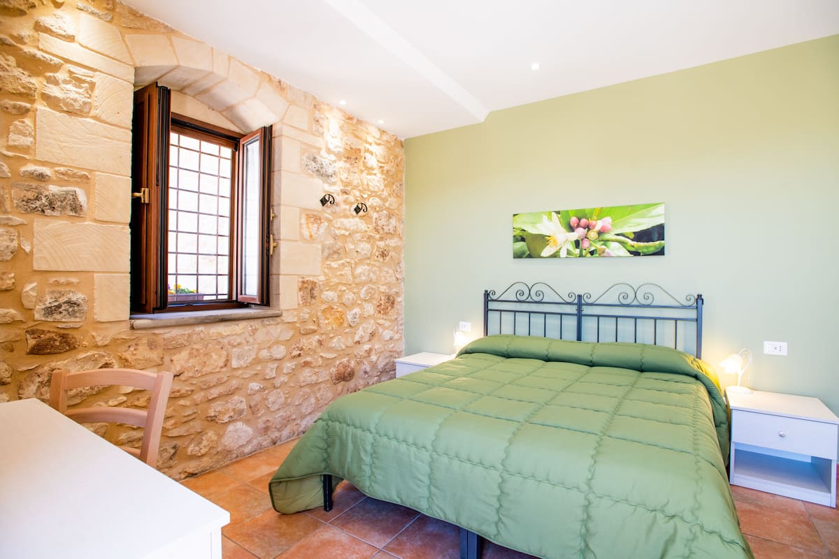 A bedroom with a stone wall, a bed with a green bedspread, a wooden chair, and a nightstand with a lamp.