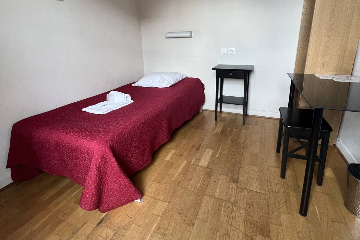 A small, single-bed room with a wooden floor, a desk, and a chair.