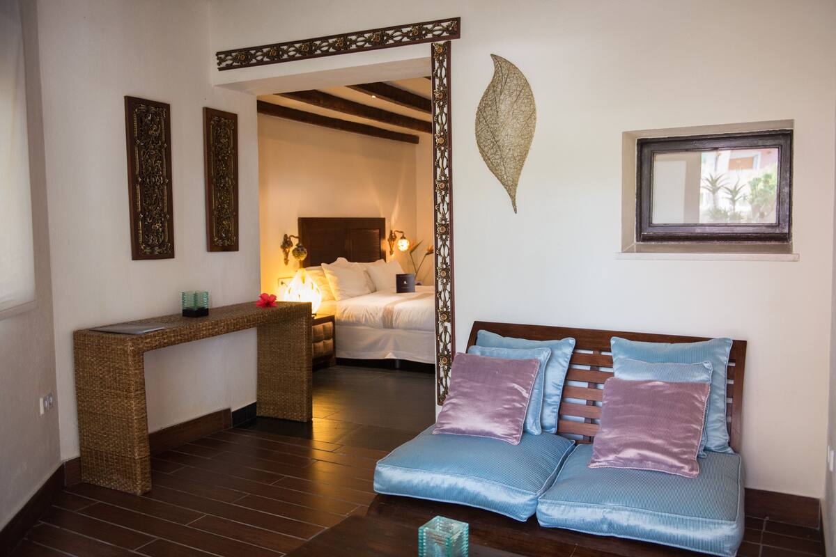 A room with a bed, a sitting area with cushions, a desk, and a decorative wall piece.