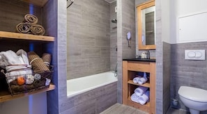Une salle de bain moderne équipée d’une baignoire, d’une douche et de toilettes. On y trouve des étagères avec des serviettes et des articles de toilette.