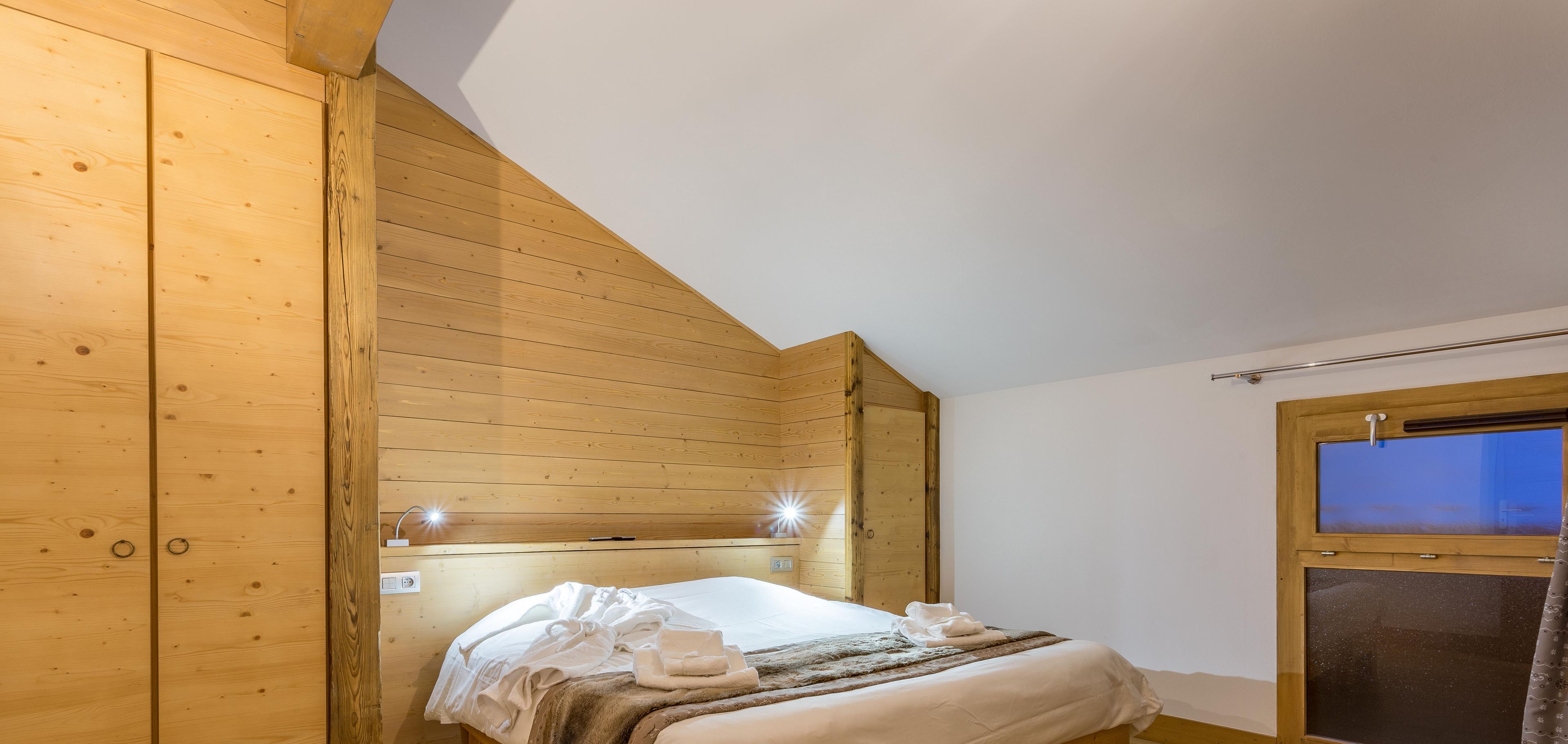 A wooden-paneled bedroom with a sloped ceiling, a bed with white linens, and a window with a view.