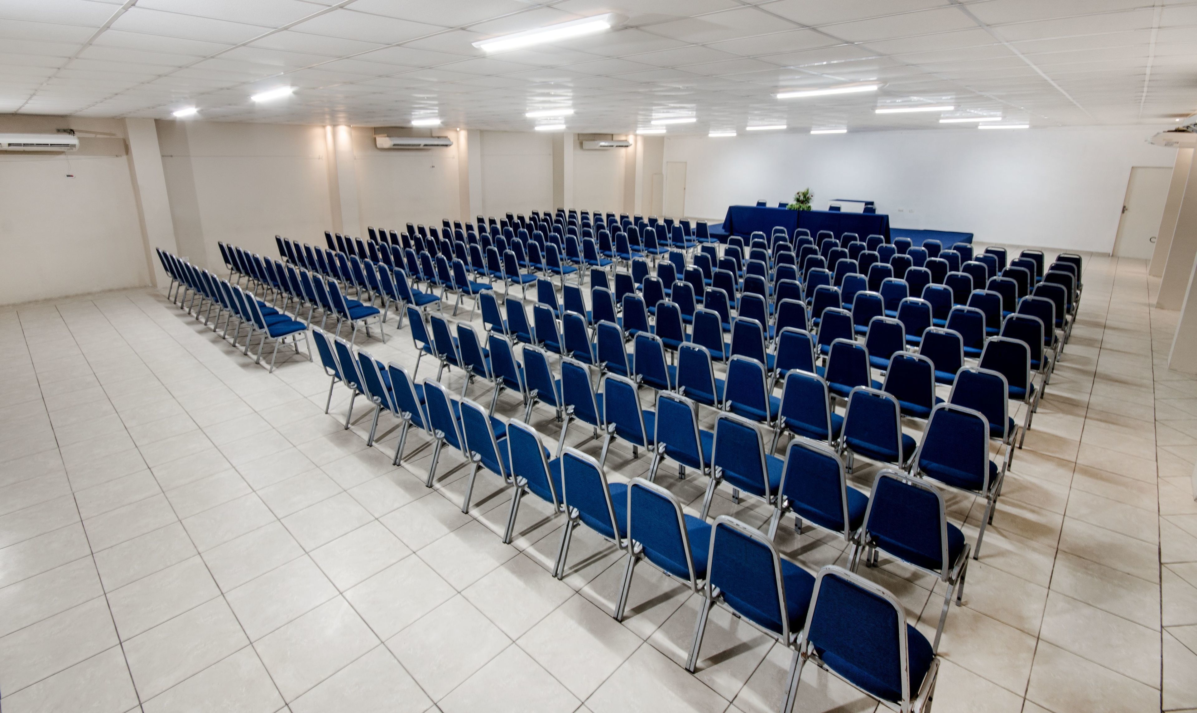 A conference room with rows of blue chairs, a podium, and air conditioning units on the ceiling.