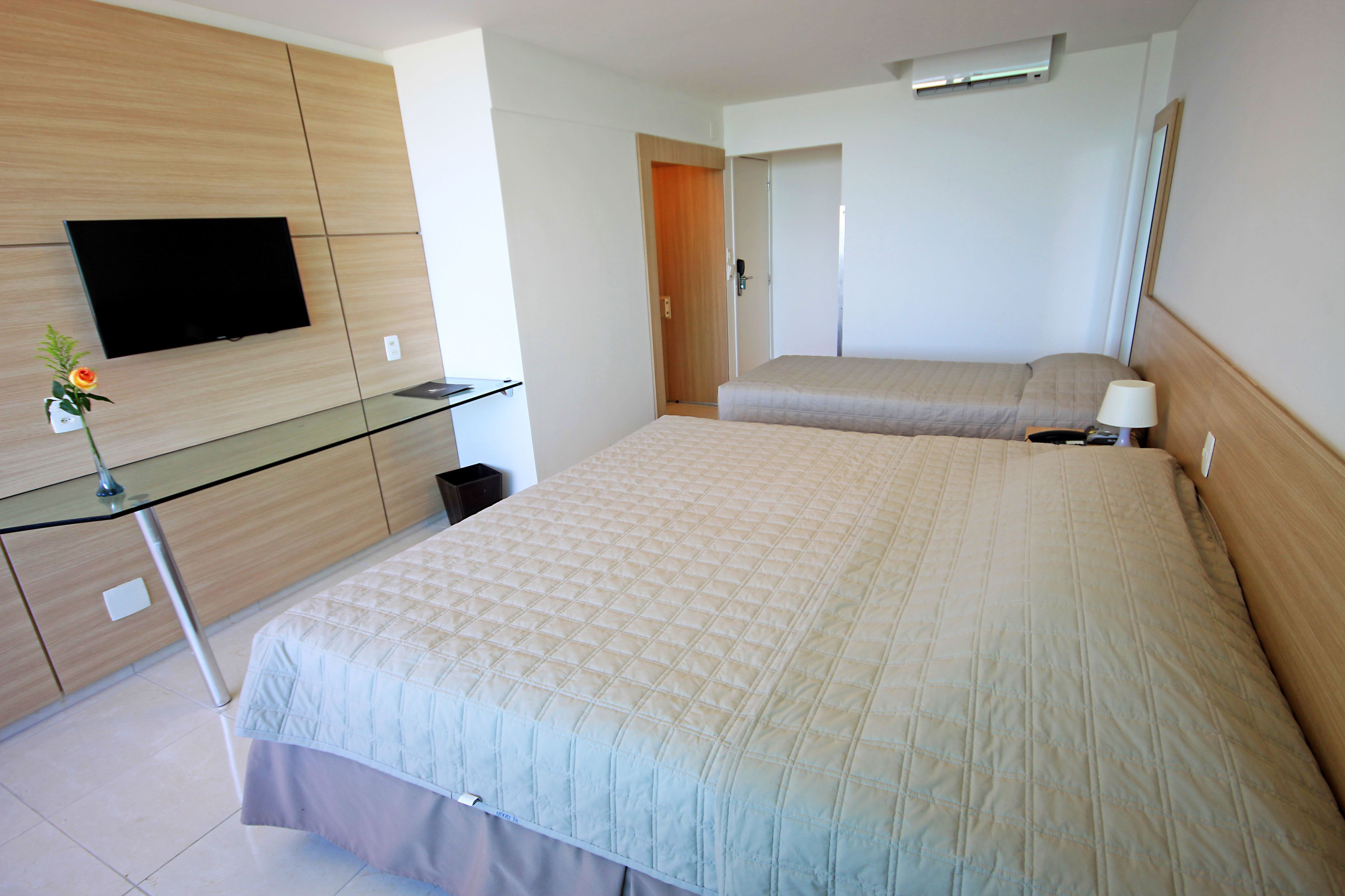 A hotel room with a bed, a TV mounted on the wall, a desk with a glass top, and a mirror on the wall.