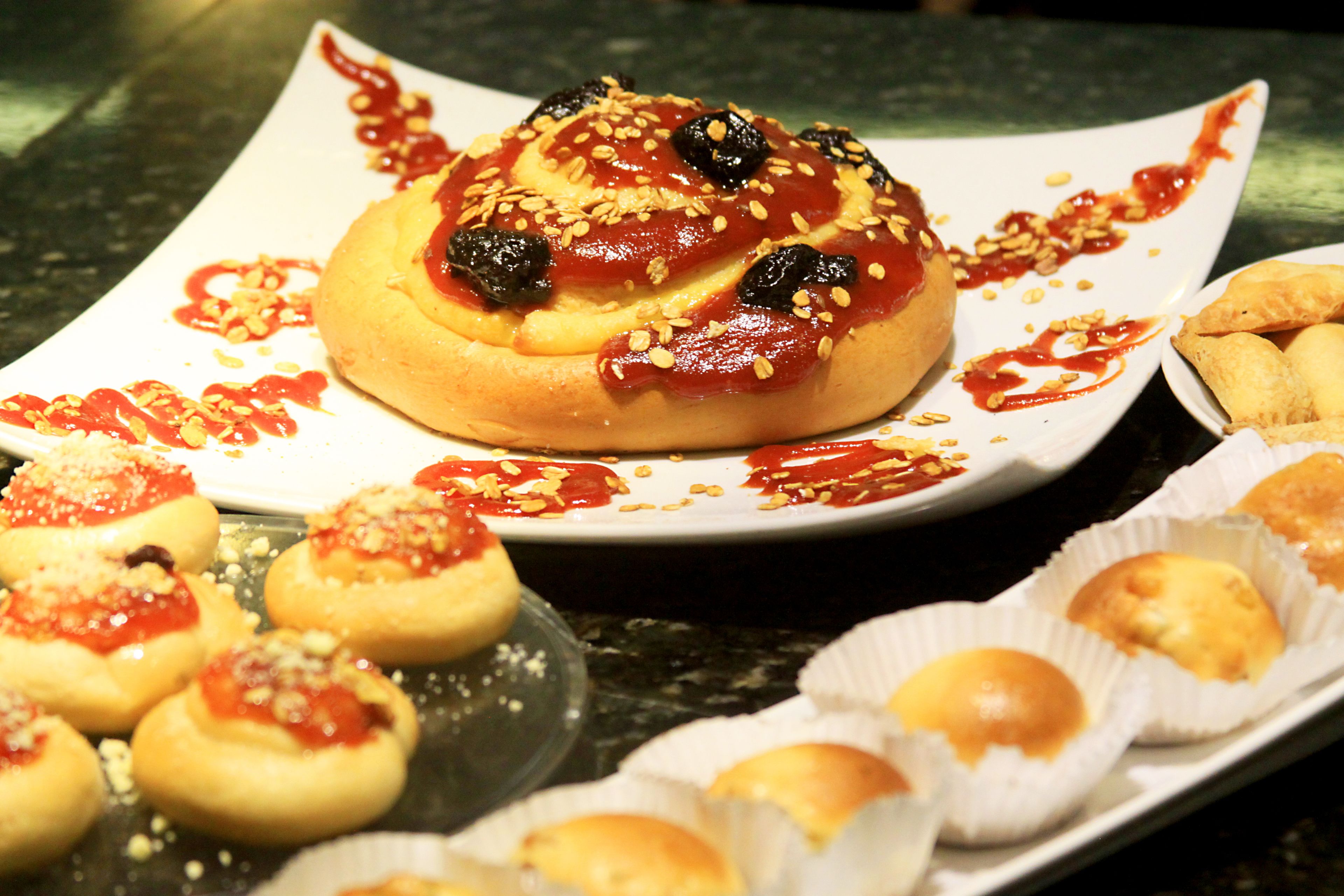 A table with various pastries, including a large bun topped with sesame seeds and a sauce, and smaller buns in paper cups.