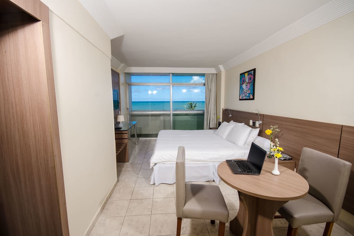 A hotel room with a bed, a desk with a laptop, a chair, and a view of the ocean.