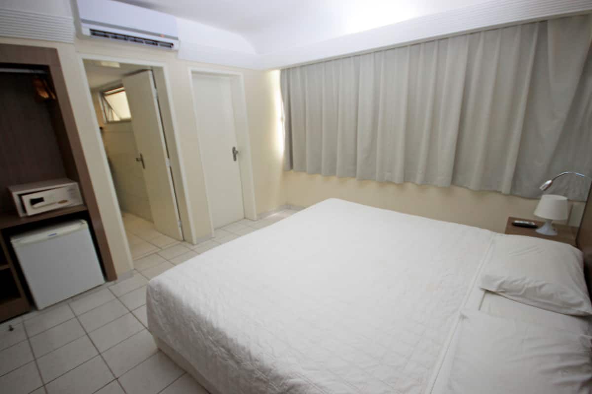 A hotel room with a bed, a nightstand, a lamp, a wardrobe, and a small refrigerator.
