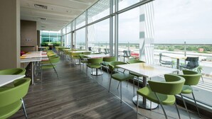 Un comedor interior moderno con sillas verdes y mesas blancas, grandes ventanales que ofrecen vistas de la ciudad.