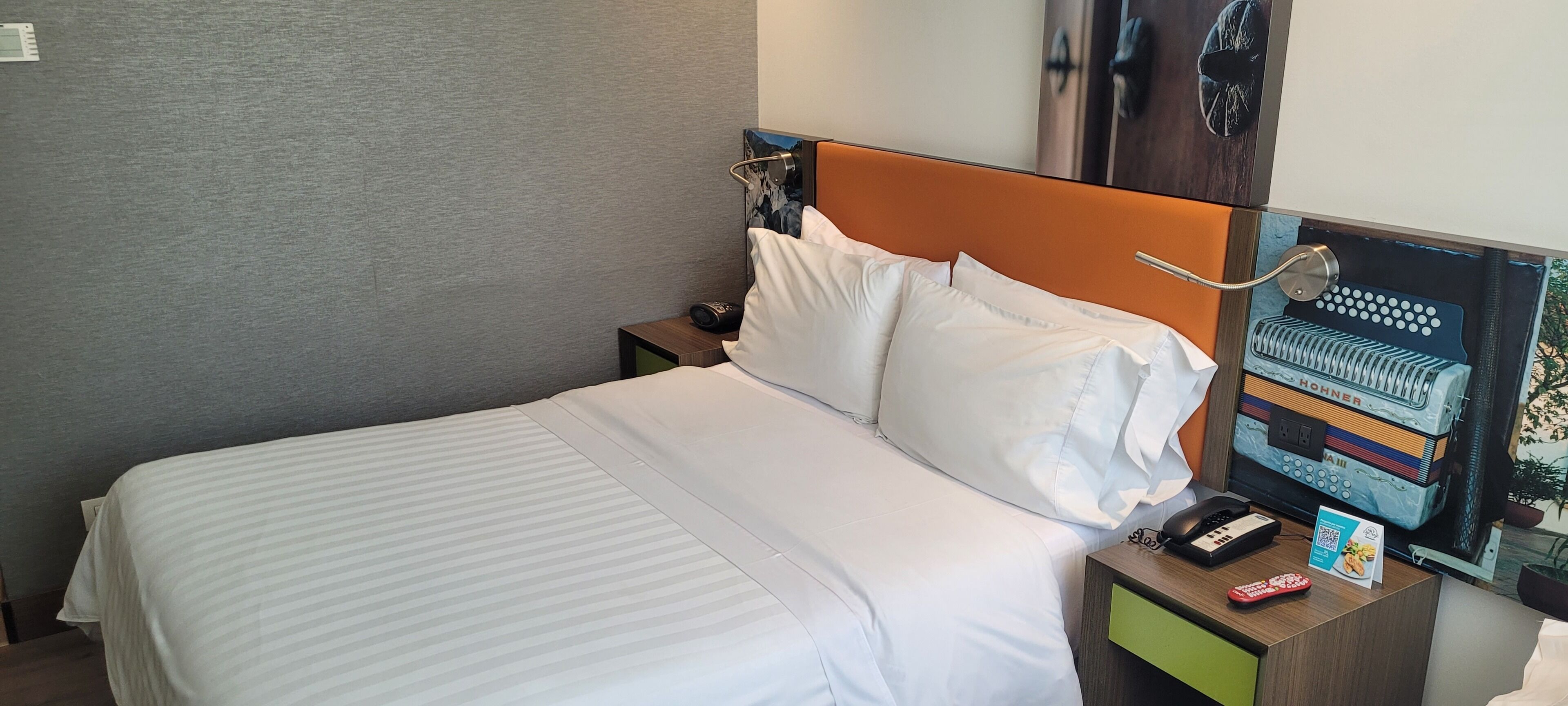 A hotel room with a bed, a nightstand, a telephone, a remote control, and a QR code sign.