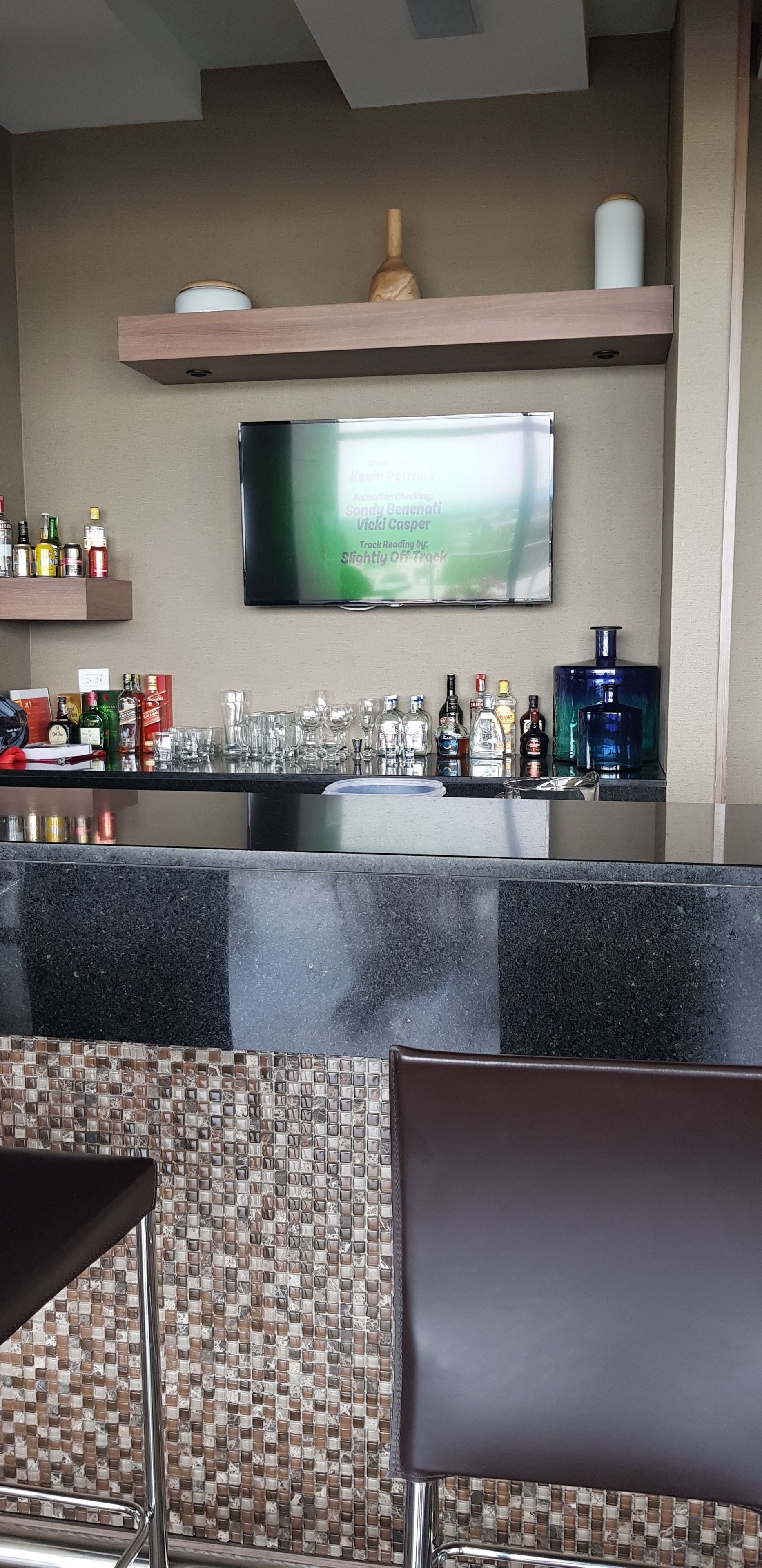Un espace bar doté d’un téléviseur à écran plat fixé au mur, d’une variété de bouteilles et de verres disposés sur le comptoir, et d’un mur recouvert de carreaux décoratifs.