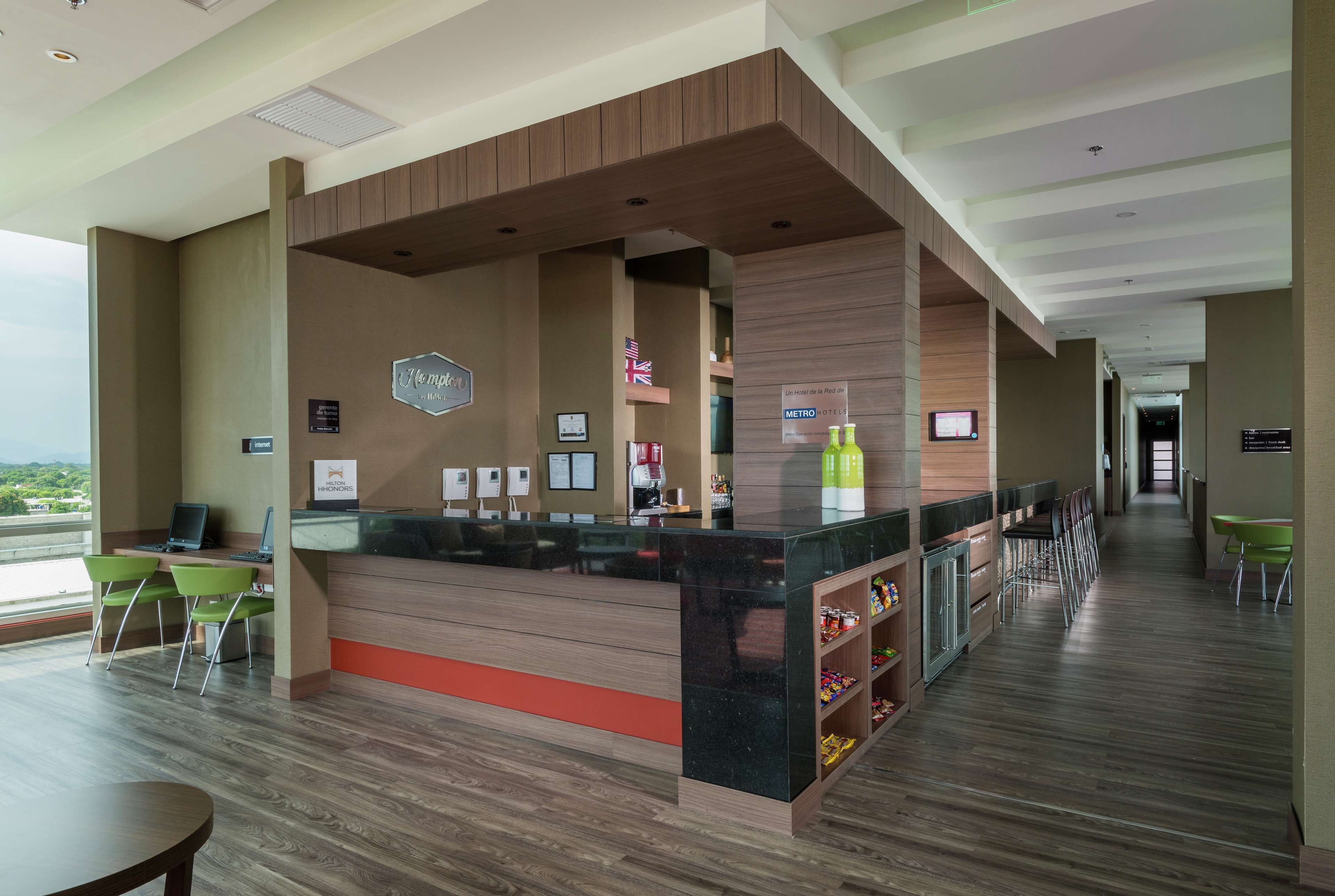 A hotel lobby with a reception desk, green chairs, a bar area, and a view of the outdoors.