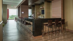 Un área de bar moderna con una barra larga y varios taburetes, un televisor montado en la pared y vistas a zonas verdes al exterior.