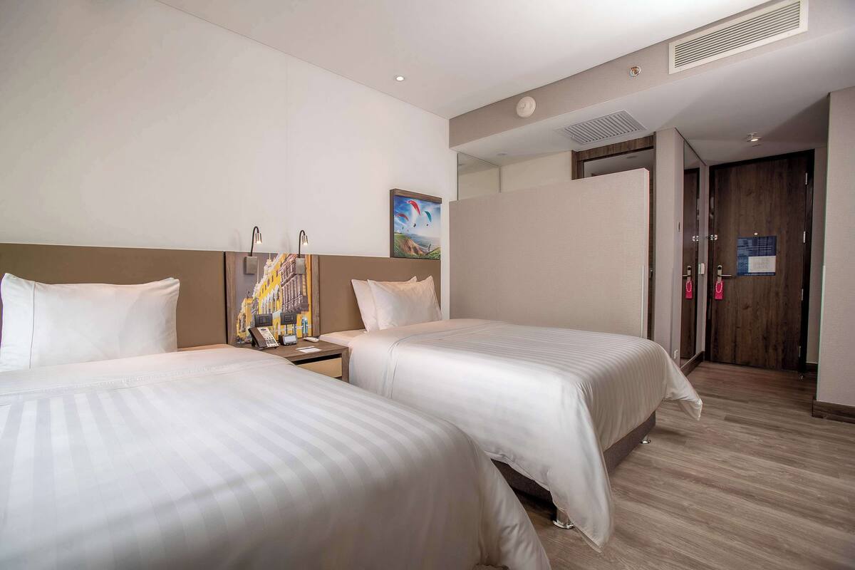 A hotel room with two beds, a nightstand, a painting on the wall, and a wooden door.