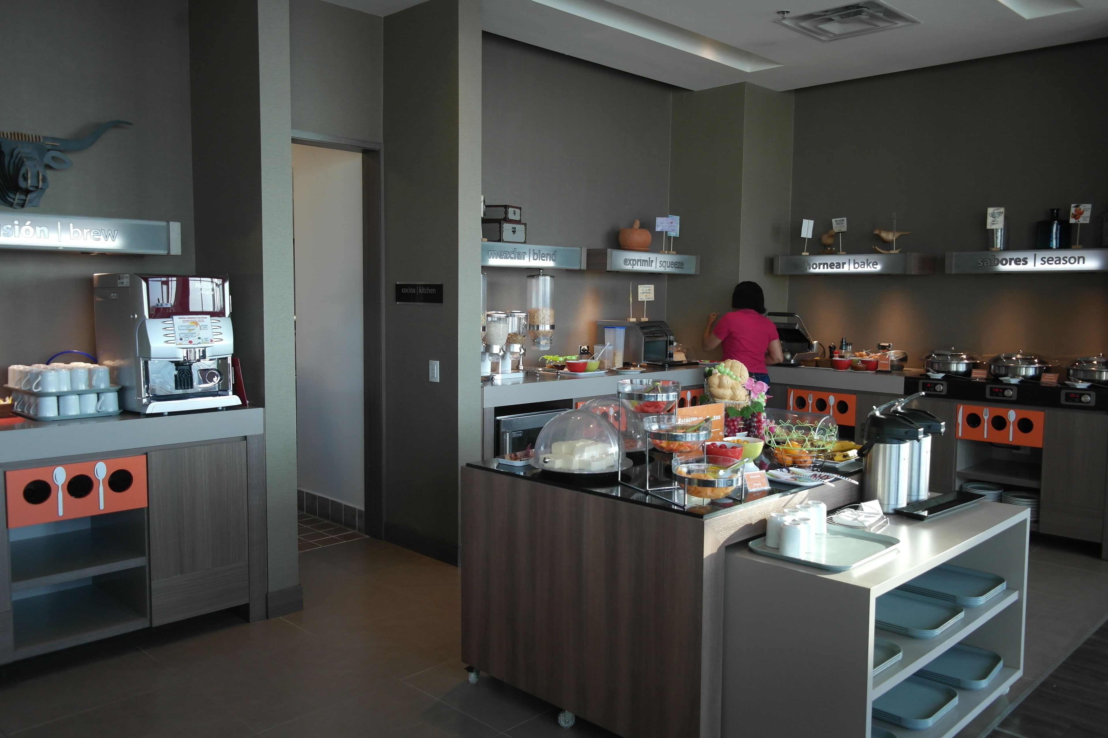 A modern kitchen area with a coffee machine, various food stations, and a person preparing food.