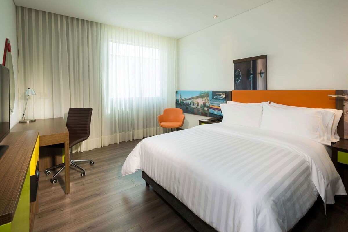 A hotel room with a bed, a desk, a chair, a window with curtains, and a painting on the wall.