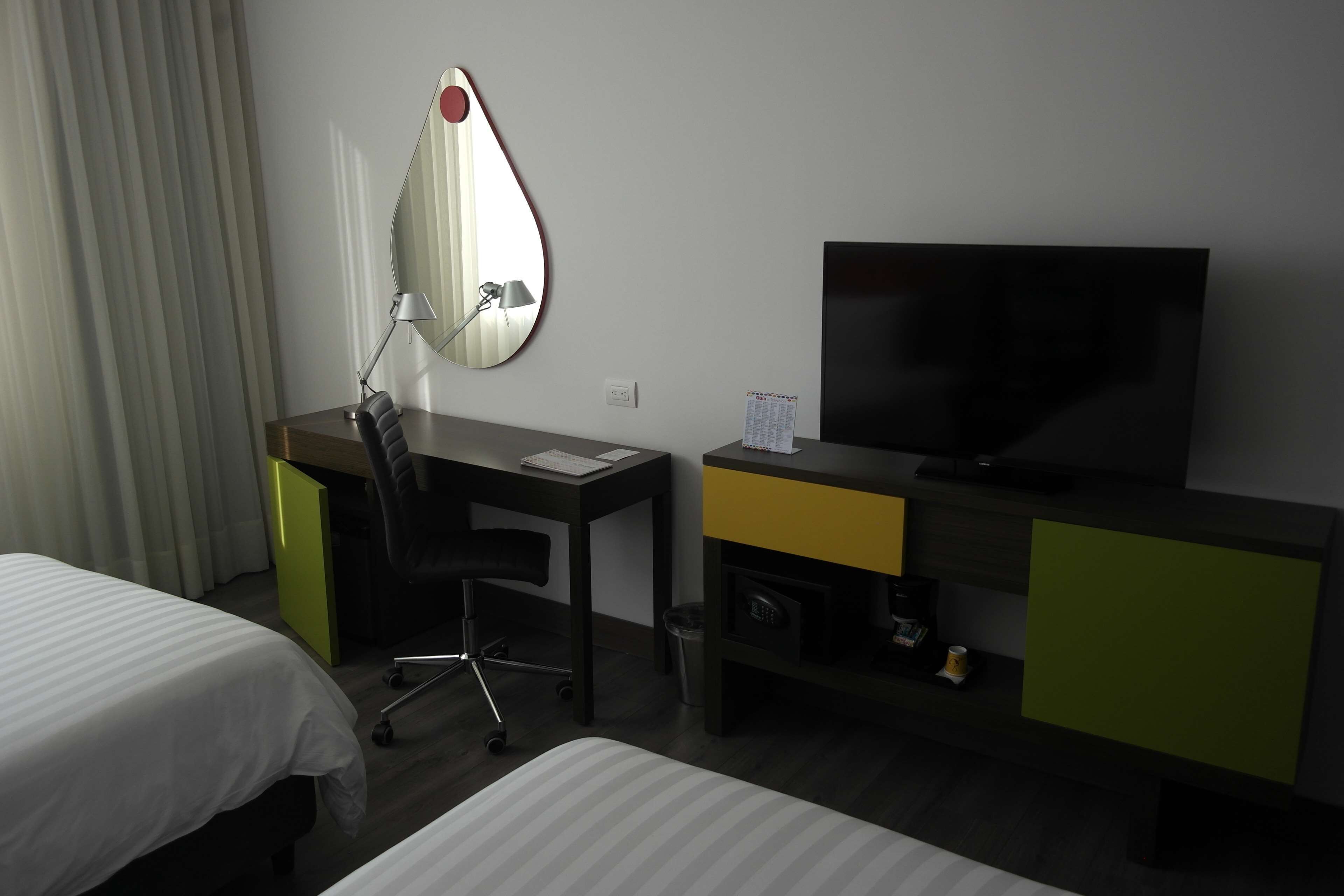 Una habitación de hotel con cama, escritorio, silla, televisor y espejo.